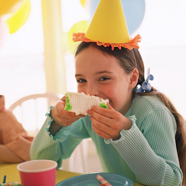 A girl smiling in a party hat eating a piece of cake with her hands.