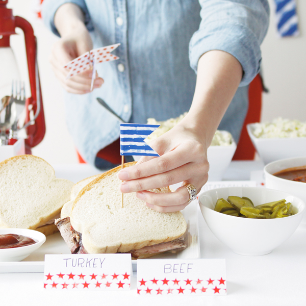A hand placing a blue striped flag food marker into a sandwich, with tented food labels marked