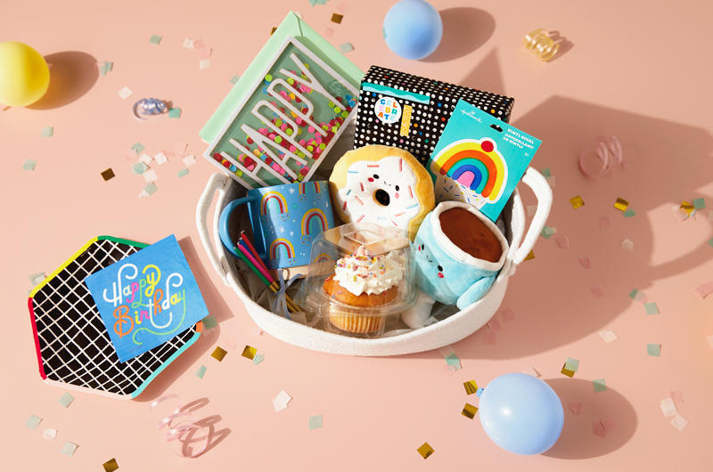 A birthday care package for a coworker or friend that includes a Coffee and Donut Better Together magnetic plush set, a light blue coffee mug with rainbows on it, a rainbow cupcake vinyl decal, tall, skinny candles, a cupcake with white frosting and rainbow-colored sprinkles, a birthday card with the word 